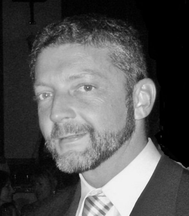 Black and white headshot of a man with short dark hair, a beard, and a mustache. He is wearing a suit and tie, looking slightly to the right.
